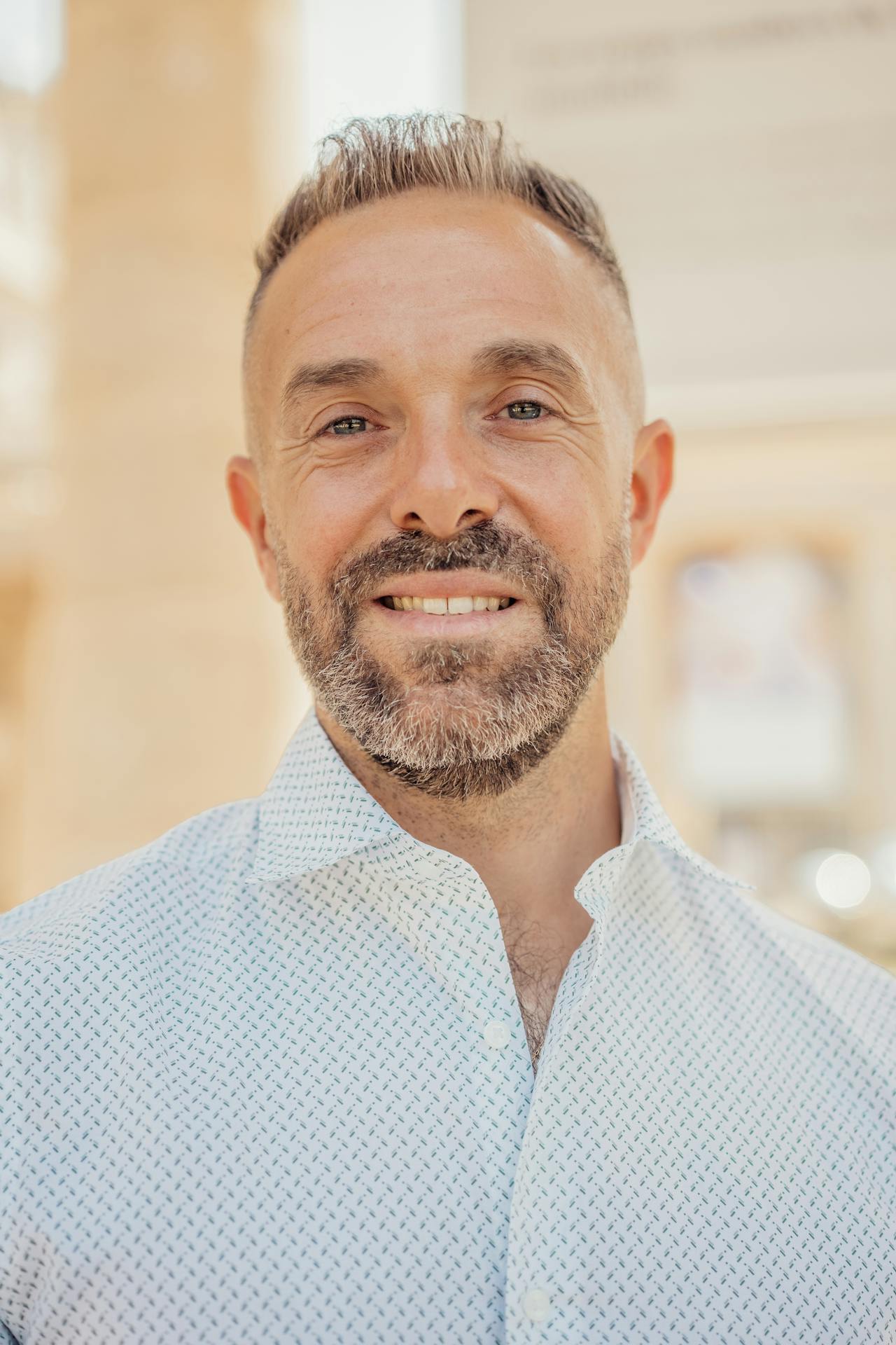 Smiling middle-aged man with short hair and beard wearing a white patterned button-up shirt.