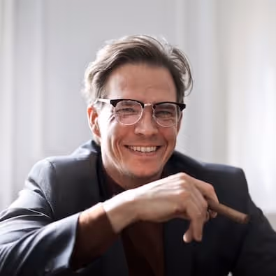 Smiling man wearing glasses and a dark blazer holding a cigar indoors.