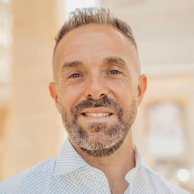 Smiling man with short hair and beard wearing a white patterned shirt against a softly blurred background.