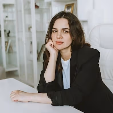 Woman with long dark hair wearing a black blazer resting her chin on her hand at a white desk in a modern room.