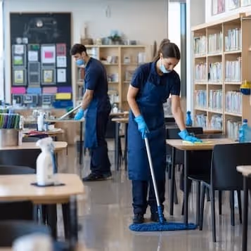 Two people wearing masks and gloves cleaning tables and floors in a library or classroom setting.