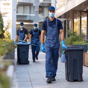 Three sanitation workers wearing masks and gloves, pushing trash bins outdoors on a paved walkway.