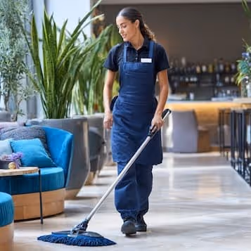Woman in navy uniform mopping floor in a modern hotel lobby with blue chairs and large green plants.