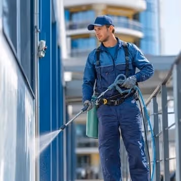 Man in blue uniform using a pressure washer to clean a building exterior.