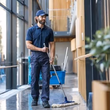 A man wearing a navy uniform mopping the floor in a modern building hallway with large windows.