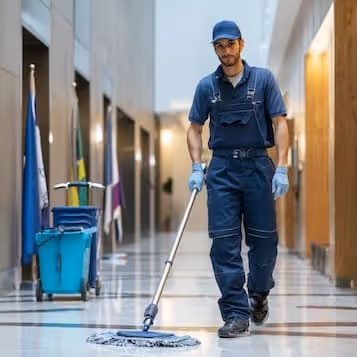 Janitor wearing blue uniform and gloves mopping floor in a building corridor.