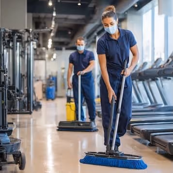 Two people wearing masks cleaning the floor with brooms inside a gym near treadmills.