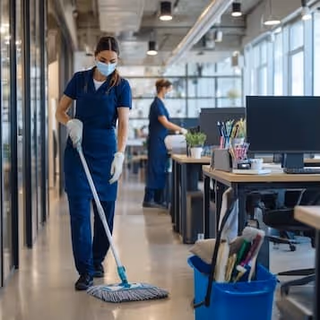 Two janitors wearing masks and gloves cleaning a modern office, one mopping the floor and the other wiping a desk.