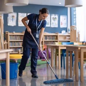 A woman in an apron mopping the floor in a classroom with colorful chairs and educational posters on the walls.