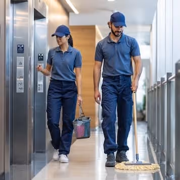 Two janitors in blue uniforms walking in a hallway, one carrying cleaning supplies and the other mopping the floor.