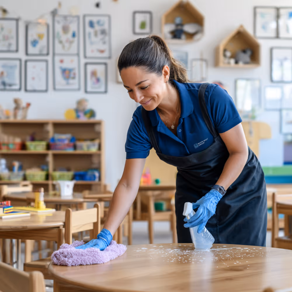 Woman wearing blue gloves and apron cleaning a wooden table with a spray bottle and cloth in a colorful classroom.