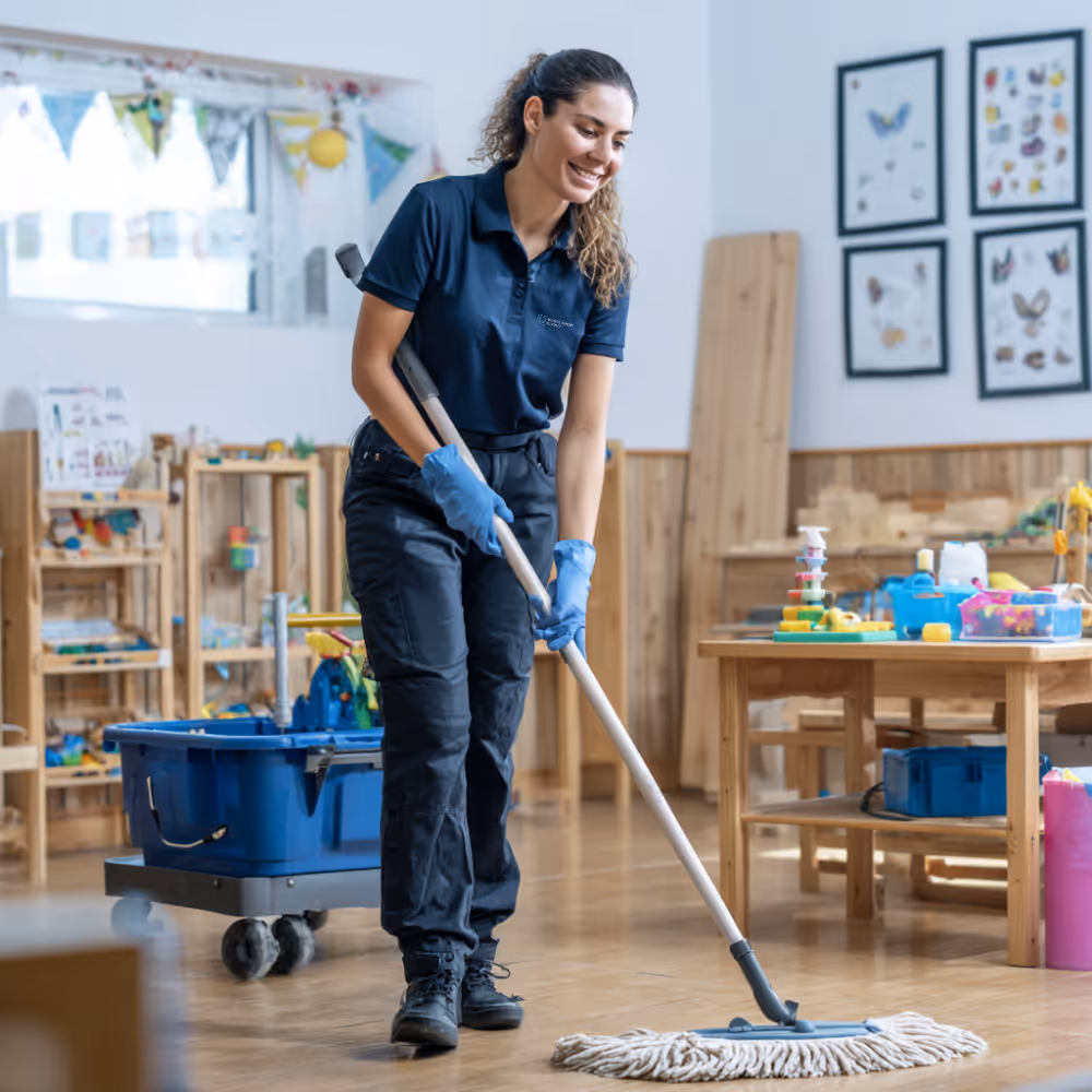 Smiling woman in blue uniform mopping the wooden floor of a bright, organized children's playroom.