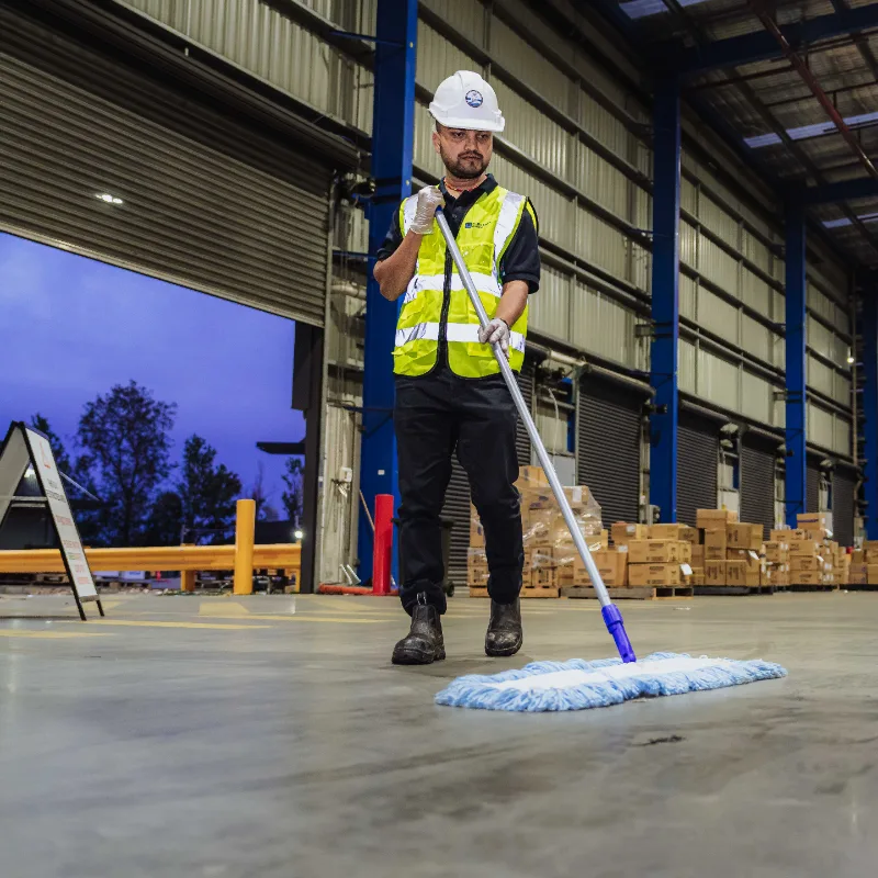 Worker wearing a white safety helmet and yellow reflective vest mopping the floor in a warehouse.
