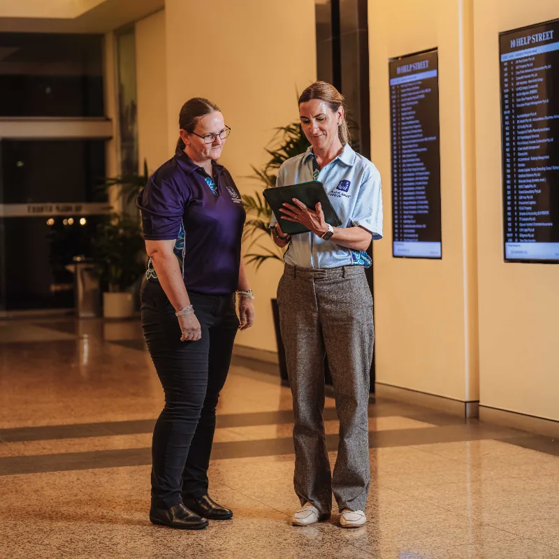 Two women standing in a hallway with digital directories, one wearing a navy polo and the other a light blue shirt, looking at a tablet.