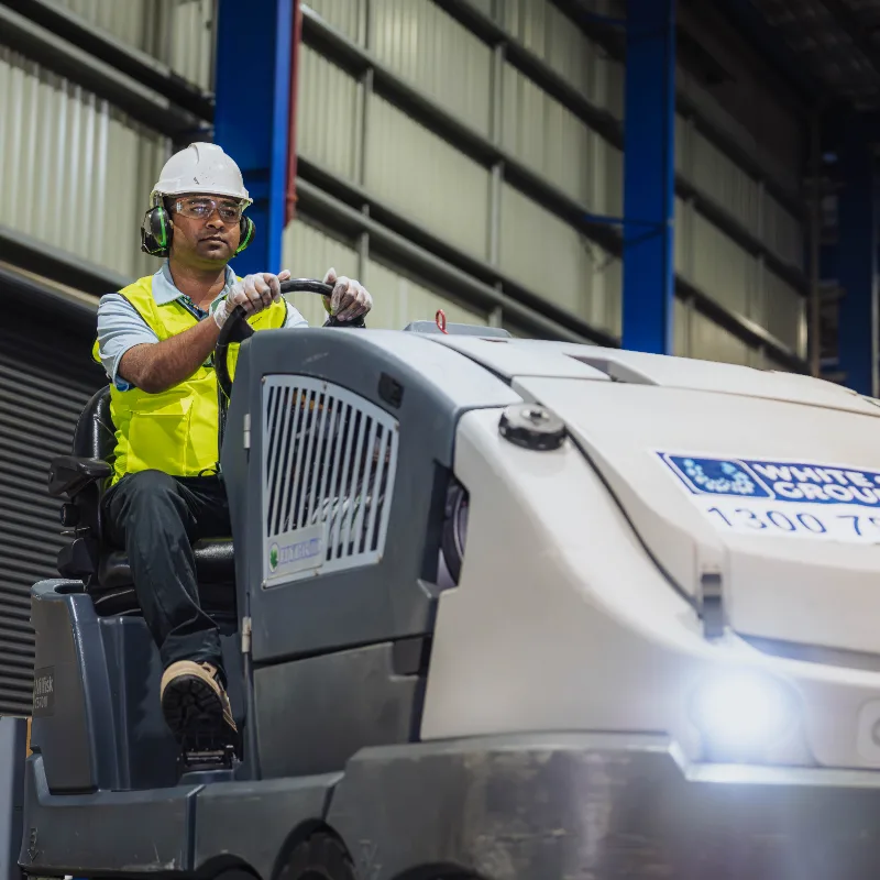 Worker wearing safety gear operating a large industrial floor cleaning machine inside a warehouse.