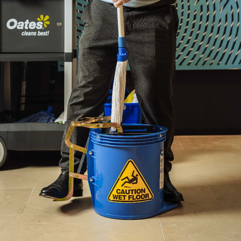 Person standing behind a blue mop bucket with a caution wet floor sign and holding a mop.