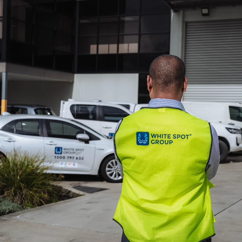 Man wearing a yellow safety vest with WHITE SPOT GROUP logo standing outdoors near parked White Spot Group vehicles.