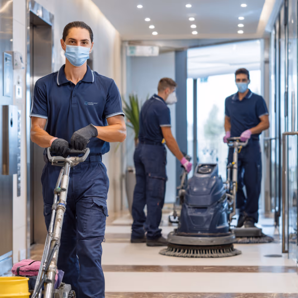 Three janitors wearing face masks and gloves operating floor cleaning machines in a building hallway.