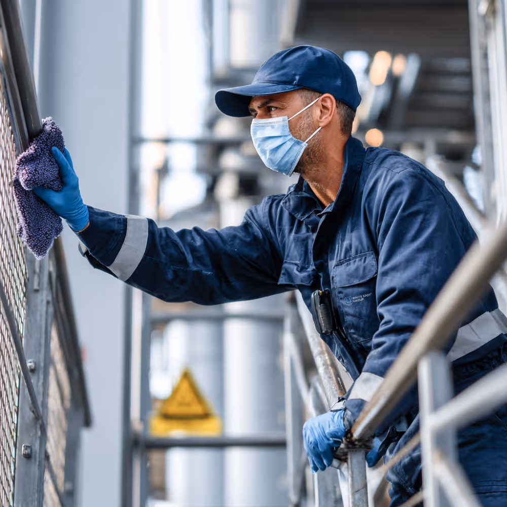 Worker in blue uniform, gloves, and face mask cleaning metal railing with a cloth in an industrial setting.