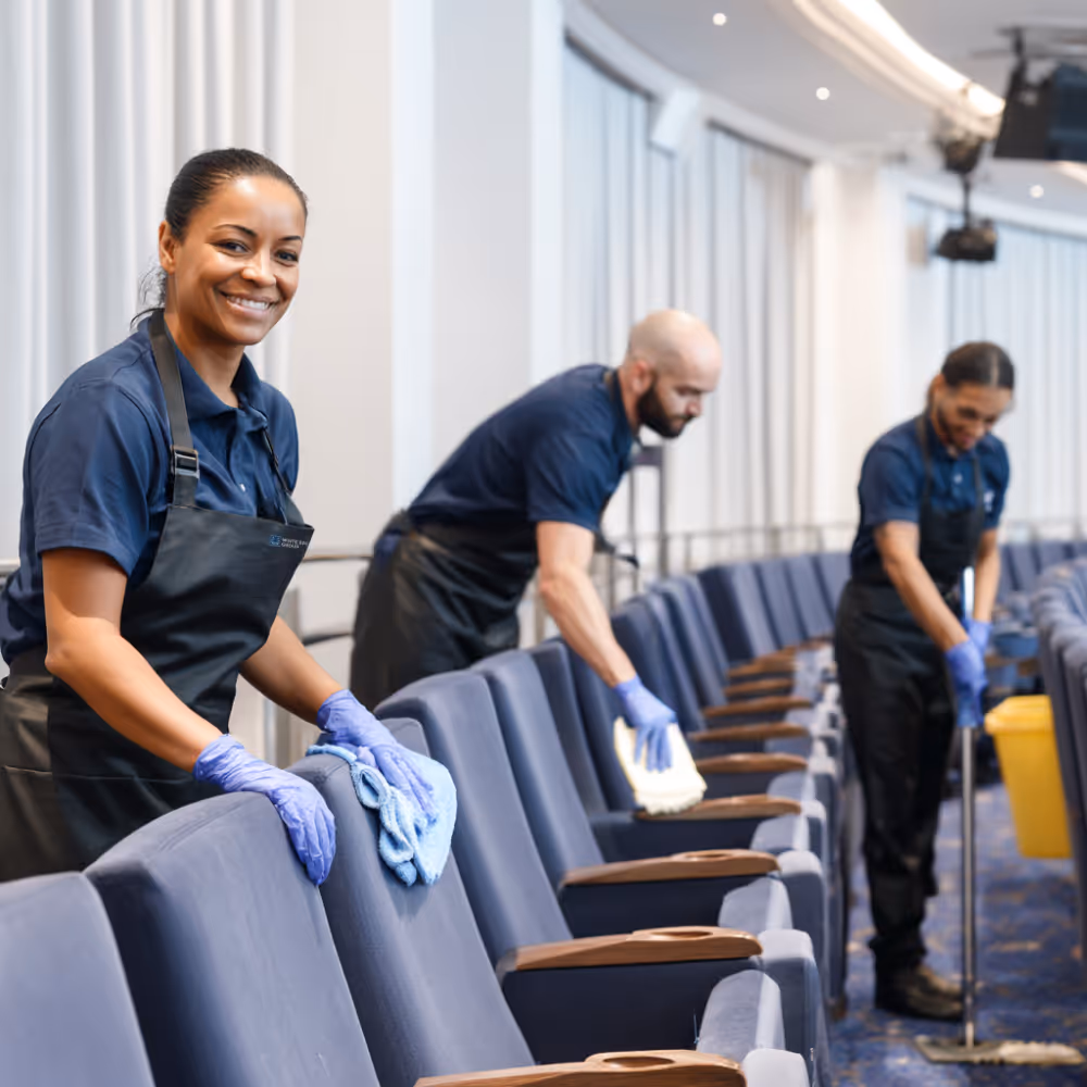 Three cleaning staff wearing gloves and aprons cleaning seats and floor in an event venue.
