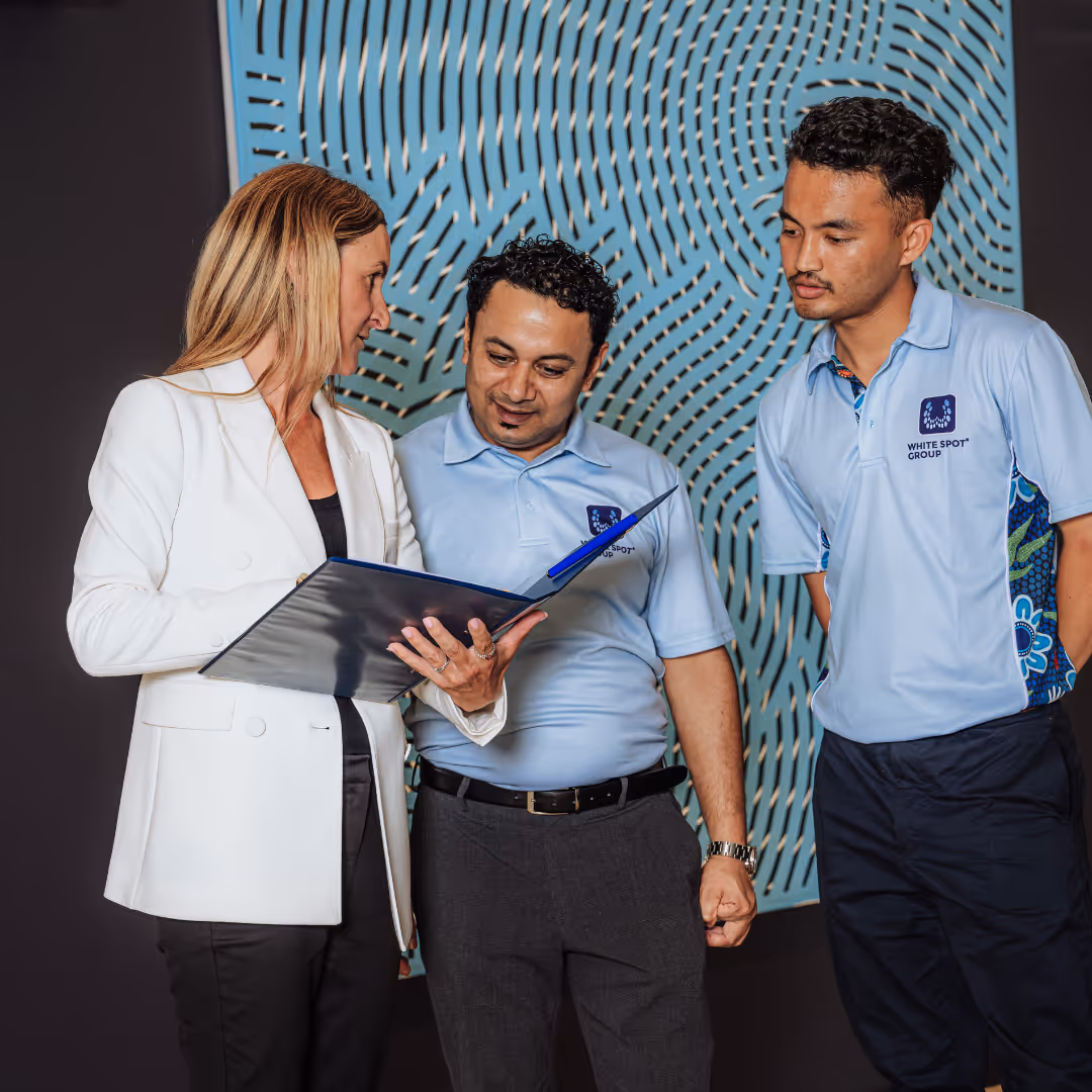 Three people, two men in light blue White Spot Group polo shirts and a woman in a white blazer, reviewing documents in a folder indoors.
