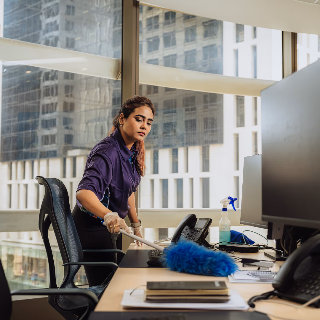 Woman in purple shirt dusting office desk near computer monitors and telephone with a blue duster.