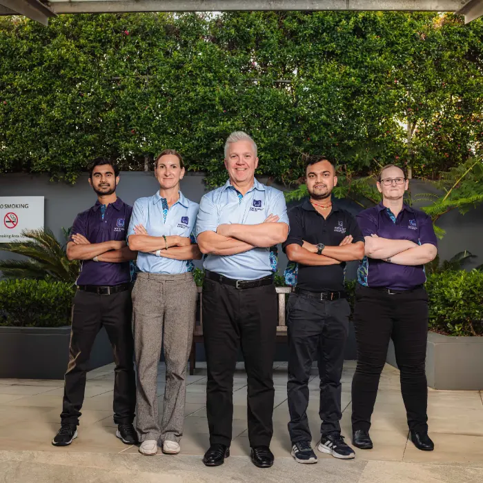 Five people standing side by side with arms crossed, wearing matching company shirts, in an outdoor area with greenery.