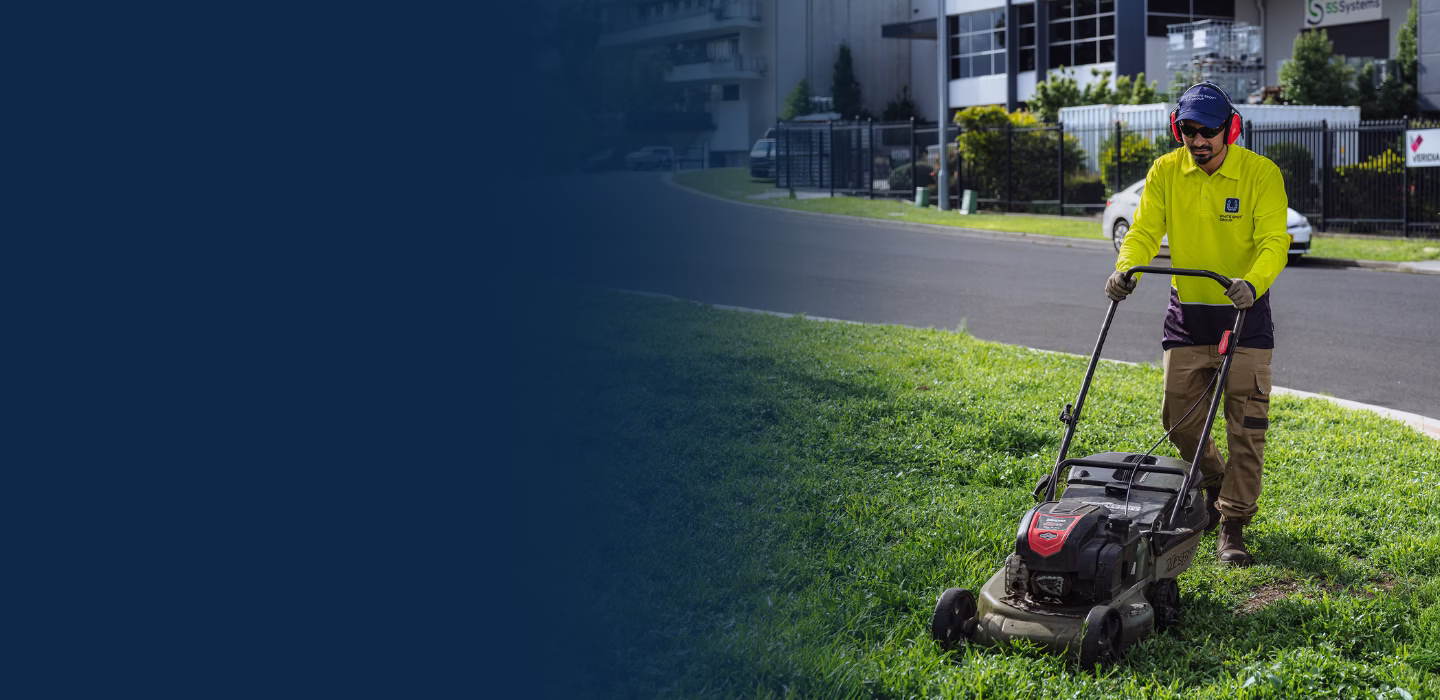 Man in safety gear mowing a green lawn with a gas-powered lawn mower near a street.
