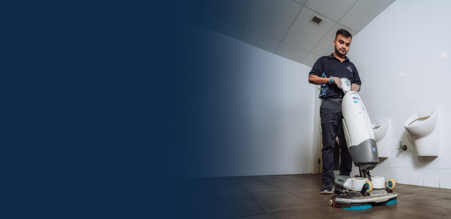 Man operating floor cleaning machine in a restroom with white tiled walls and urinals.