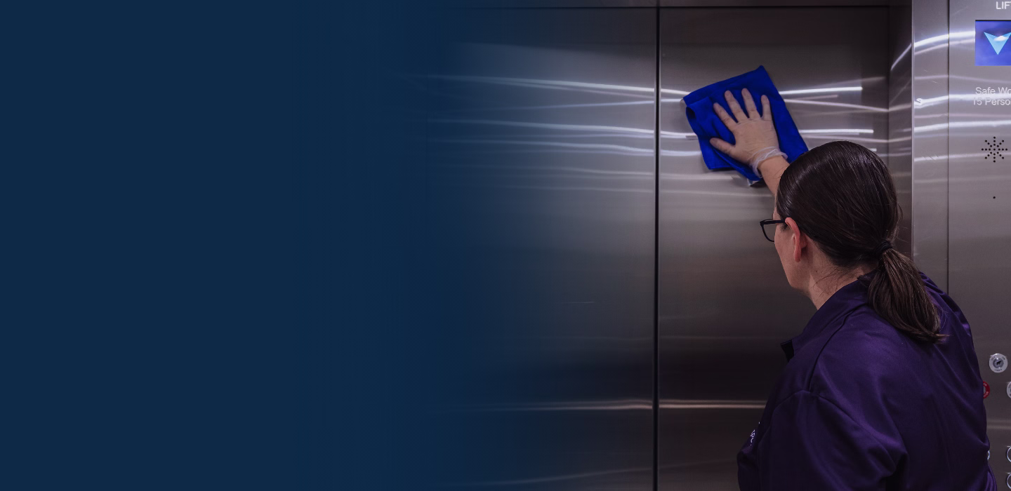 Woman wearing gloves cleaning the interior stainless steel door of an elevator with a blue cloth.
