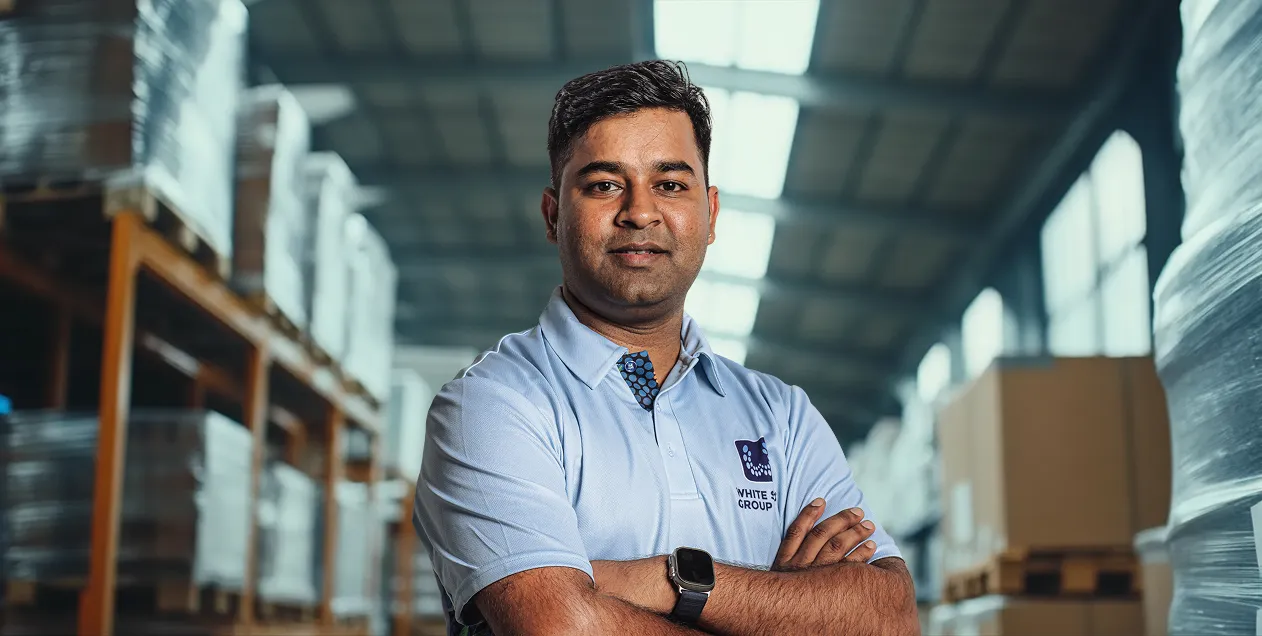 Man in a light blue shirt with White Group logo standing confidently with arms crossed in a warehouse environment.