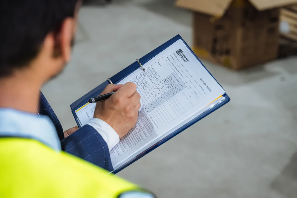 Person in a yellow safety vest holding a clipboard and marking a warehouse checklist.