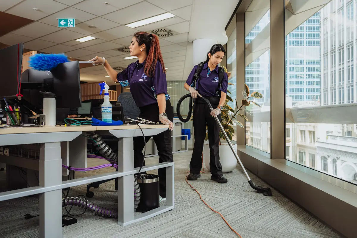 Two female cleaners in uniform dusting an office desk and vacuuming carpet near large office windows.