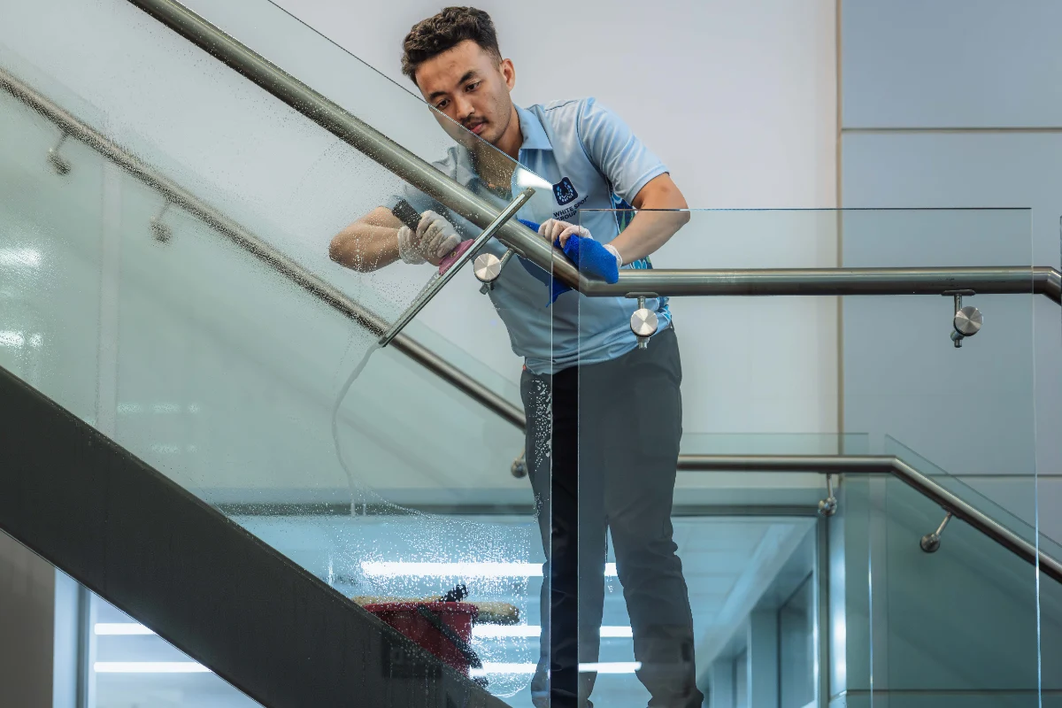 Man wearing a light blue uniform cleaning a glass railing of a staircase with a squeegee and blue cloth indoors.