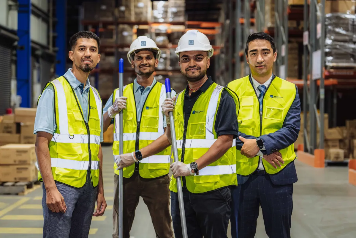 Four warehouse workers in safety vests and helmets standing in an industrial storage area with pallets and shelves.