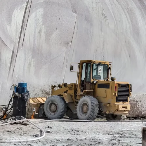Yellow mining loader vehicle parked inside a large tunnel with concrete walls and scattered equipment on dusty ground.