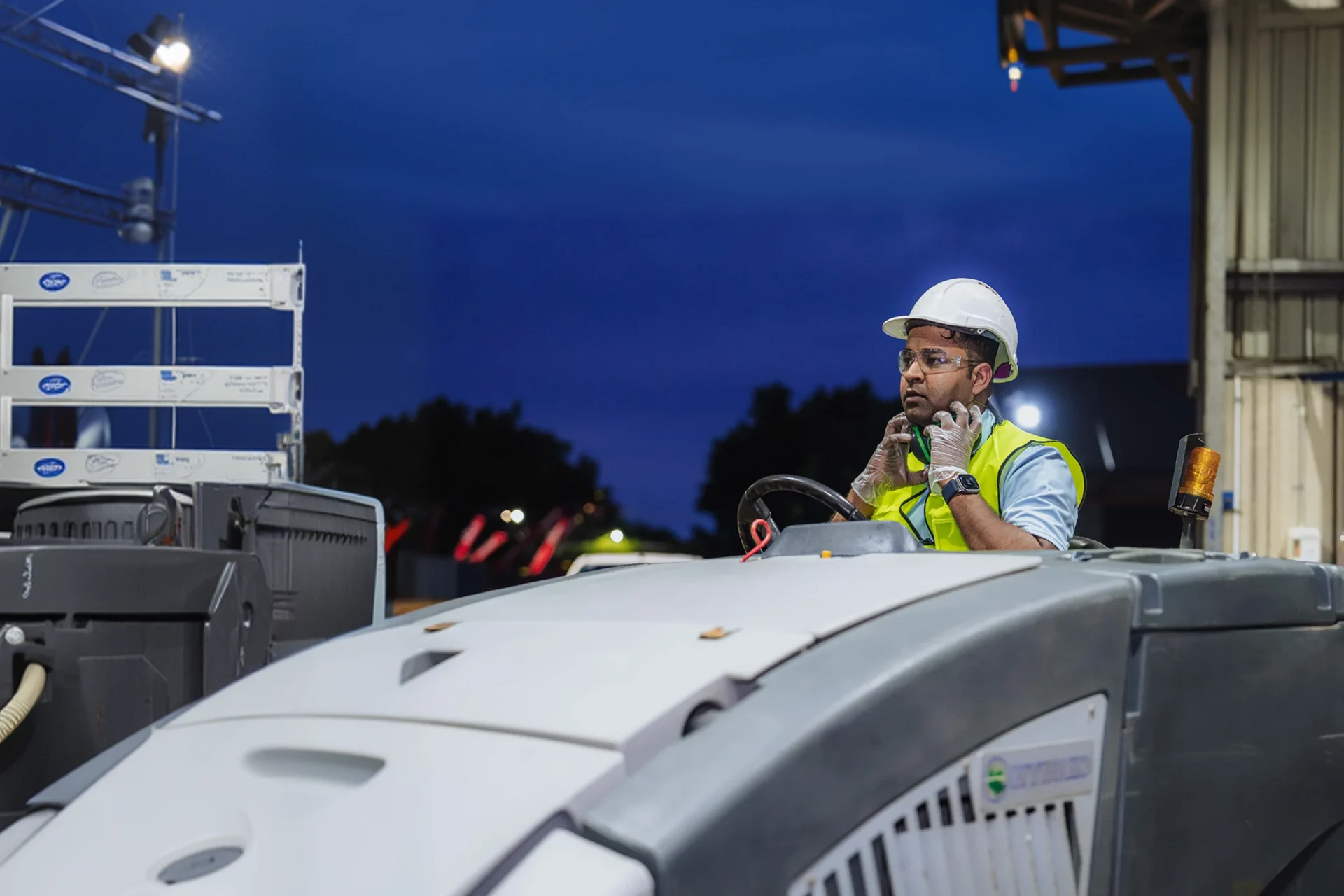 Worker wearing safety helmet, glasses, gloves, and high-visibility vest, sitting on industrial machinery in a warehouse at night.