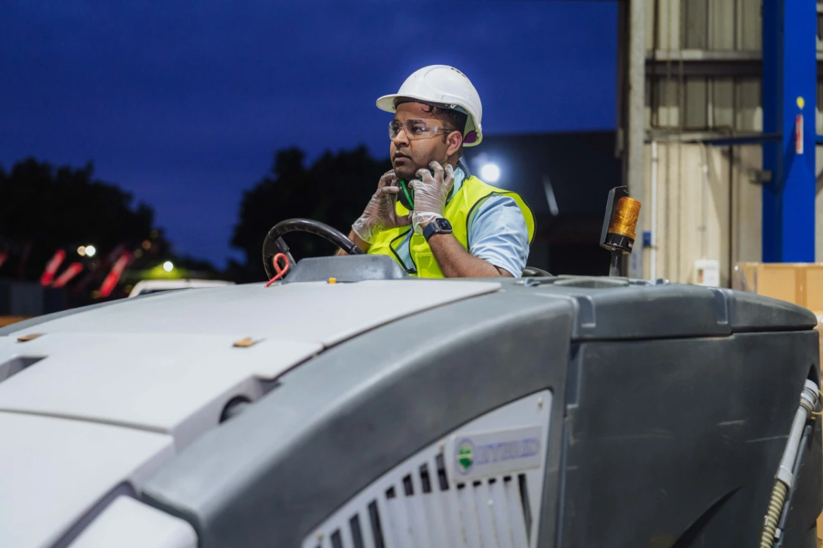 Worker wearing safety helmet, glasses, gloves, and high-visibility vest, sitting on industrial machinery in a warehouse at night.