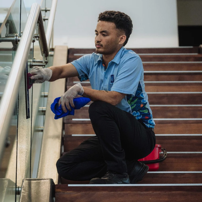 Man in blue shirt and gloves cleaning a glass railing on wooden stairs with a cloth and sponge.