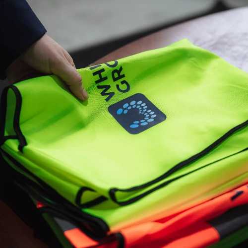Hand holding a folded bright yellow high-visibility safety vest with a black logo and text partially visible on a table with more vests underneath.