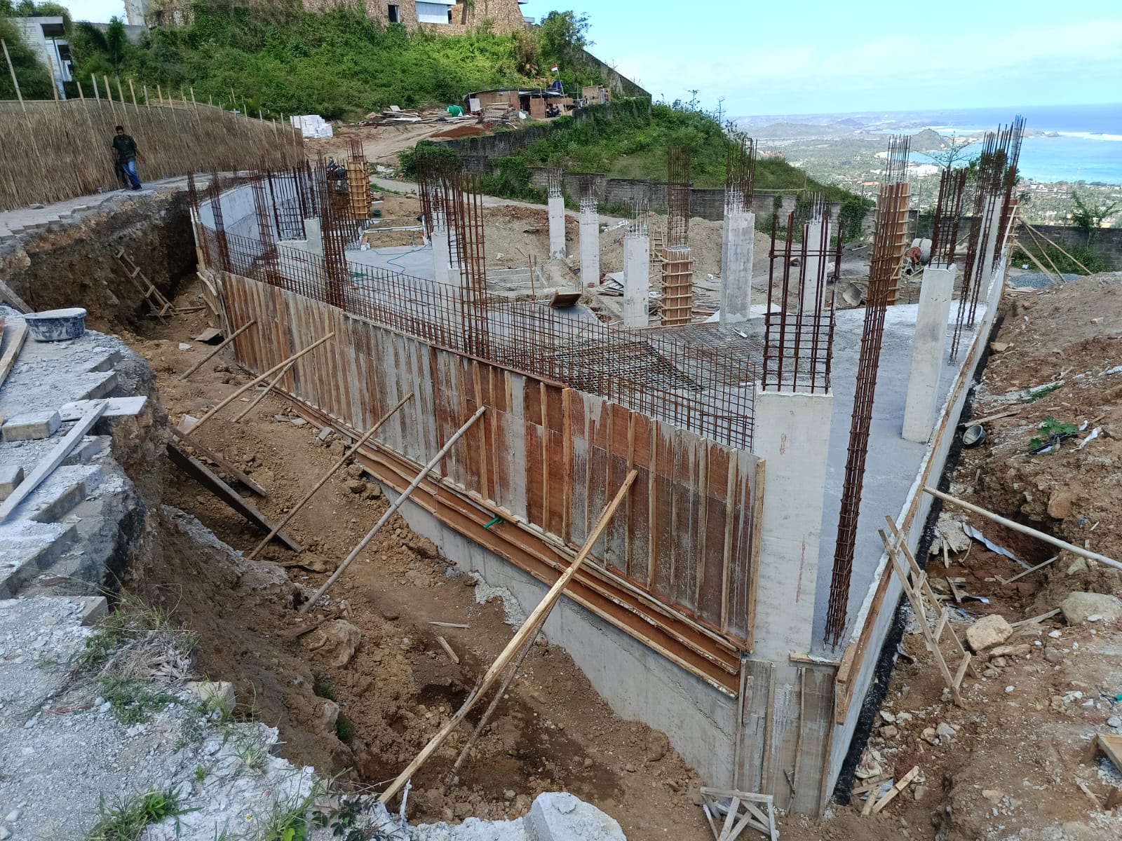 Construction site with concrete foundation, vertical steel reinforcement bars, and wooden formwork on a hillside with a distant ocean view.