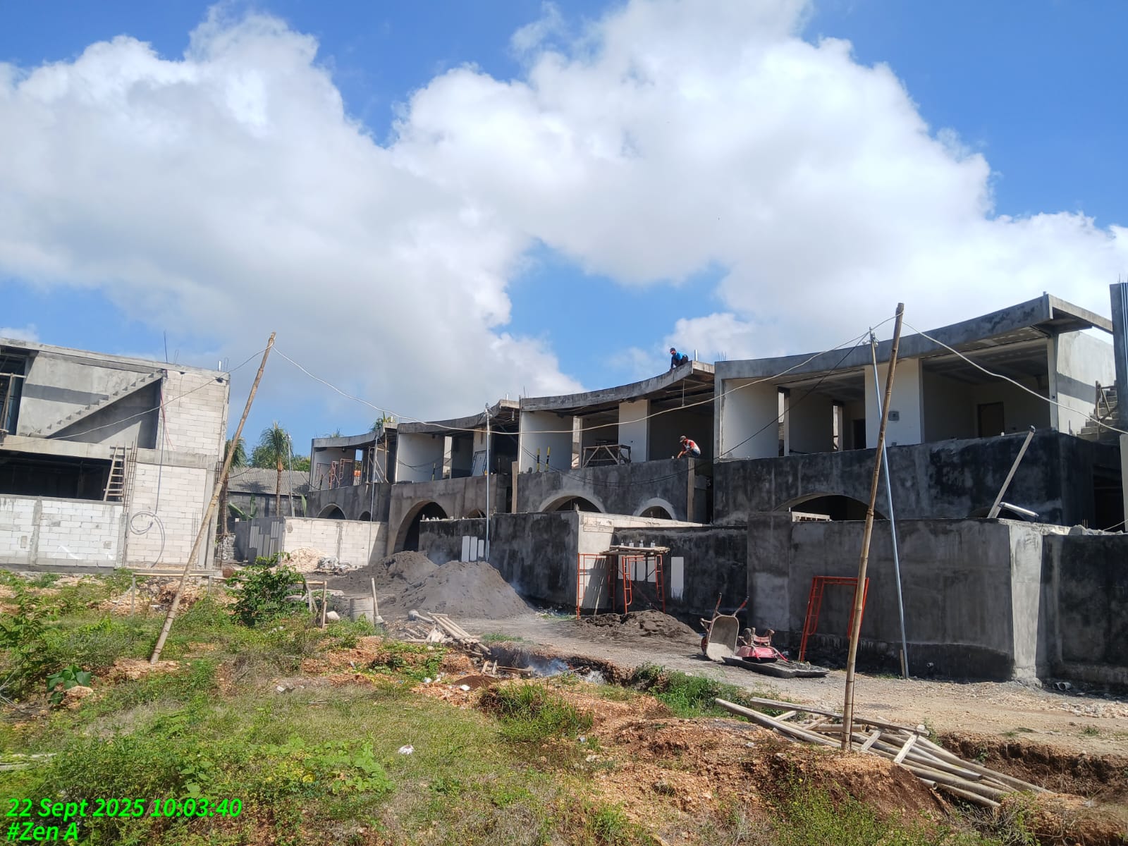 Partially constructed multi-unit building with workers on the upper level and construction materials scattered on the ground under a blue sky with clouds.