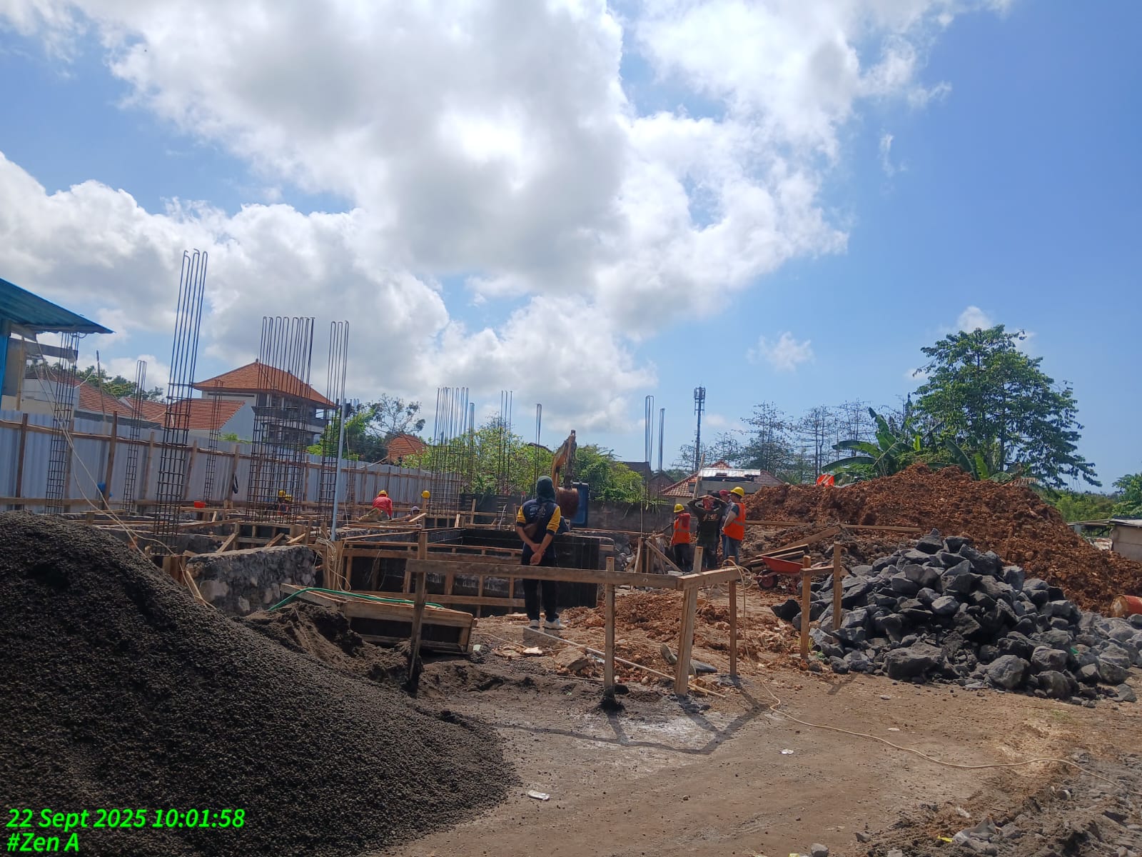 Construction site with workers, piles of soil and rocks, rebar structures, and a partly cloudy sky.