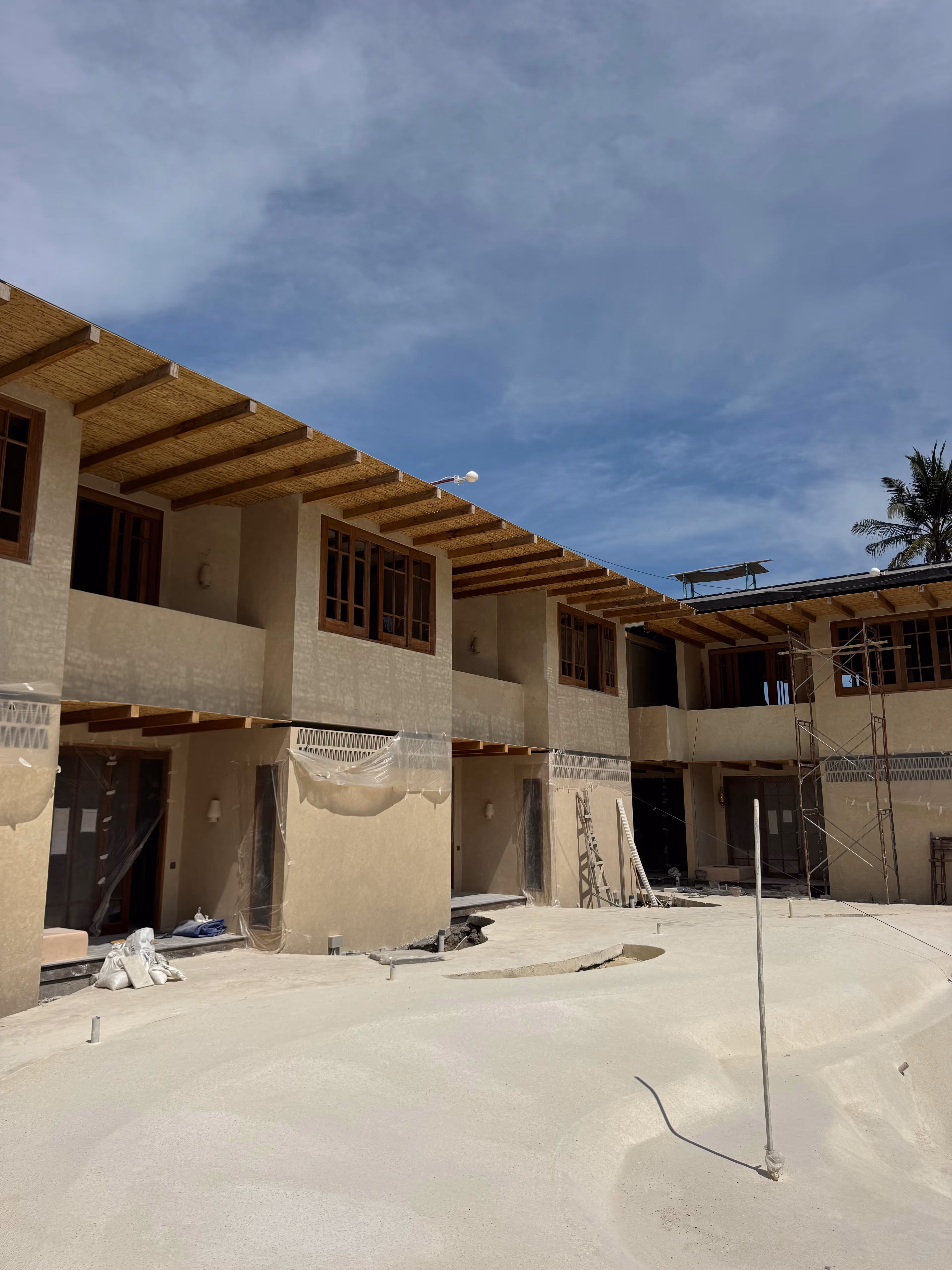 Under-construction beige two-story building with wooden window frames and a large unfinished courtyard under a partly cloudy sky.