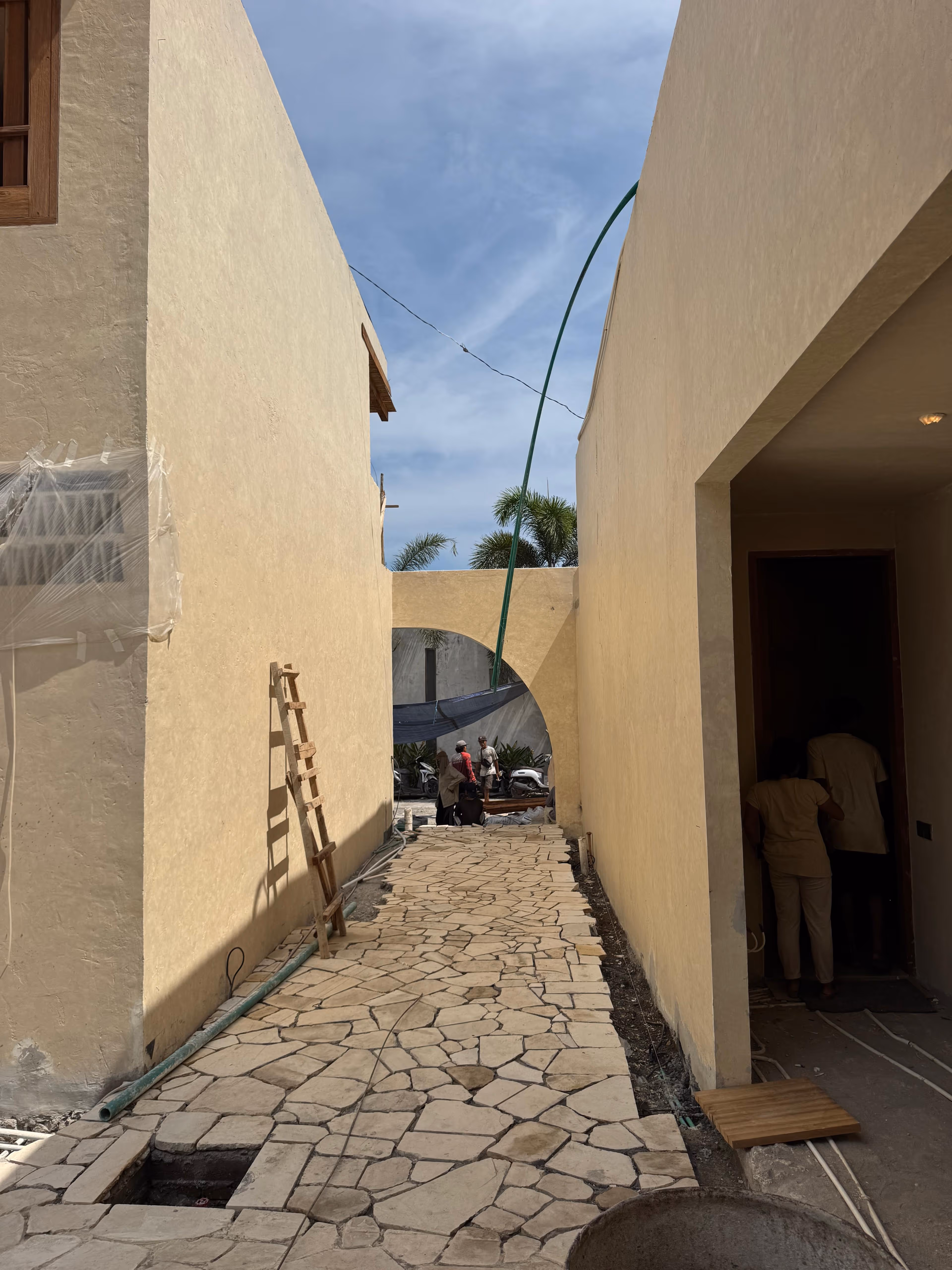 Narrow stone pathway between beige buildings with a wooden ladder, green hose, and two people standing under an archway.