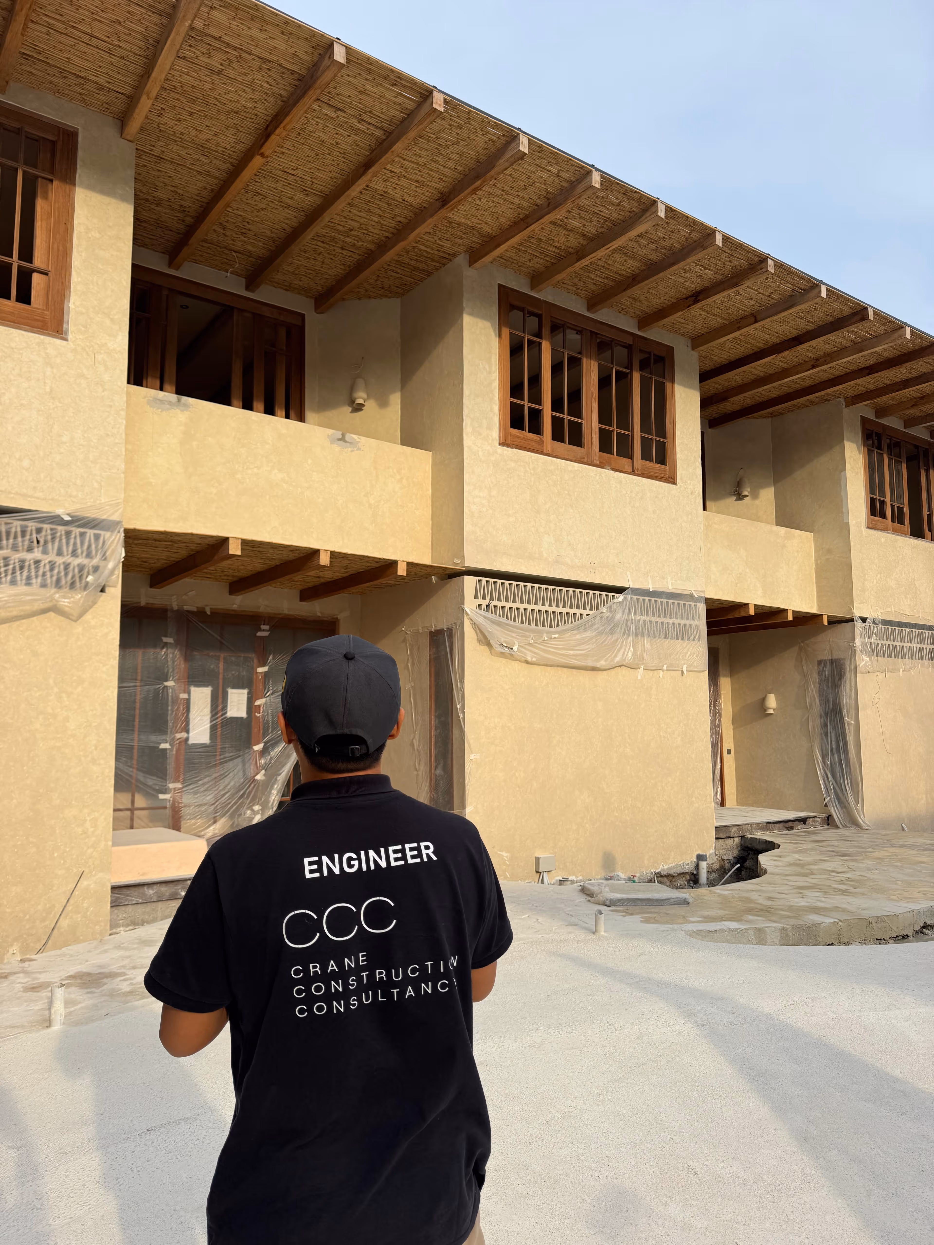 Engineer wearing a black shirt with 'CRANE CONSTRUCTION CONSULTANCY' on the back, standing in front of a beige building under construction.