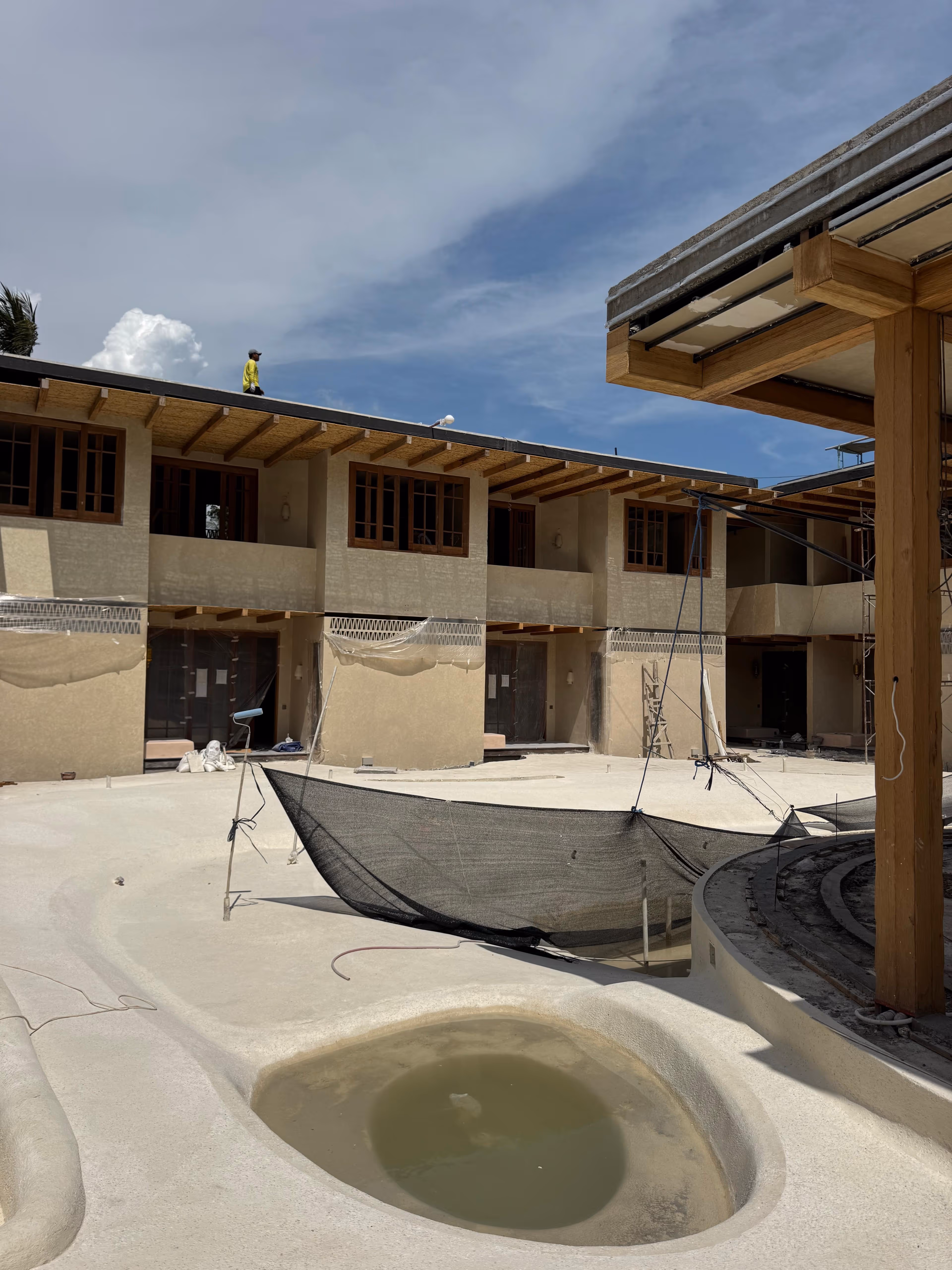 Construction site of a two-story building with unfinished pool and worker standing on the roof under a blue sky.