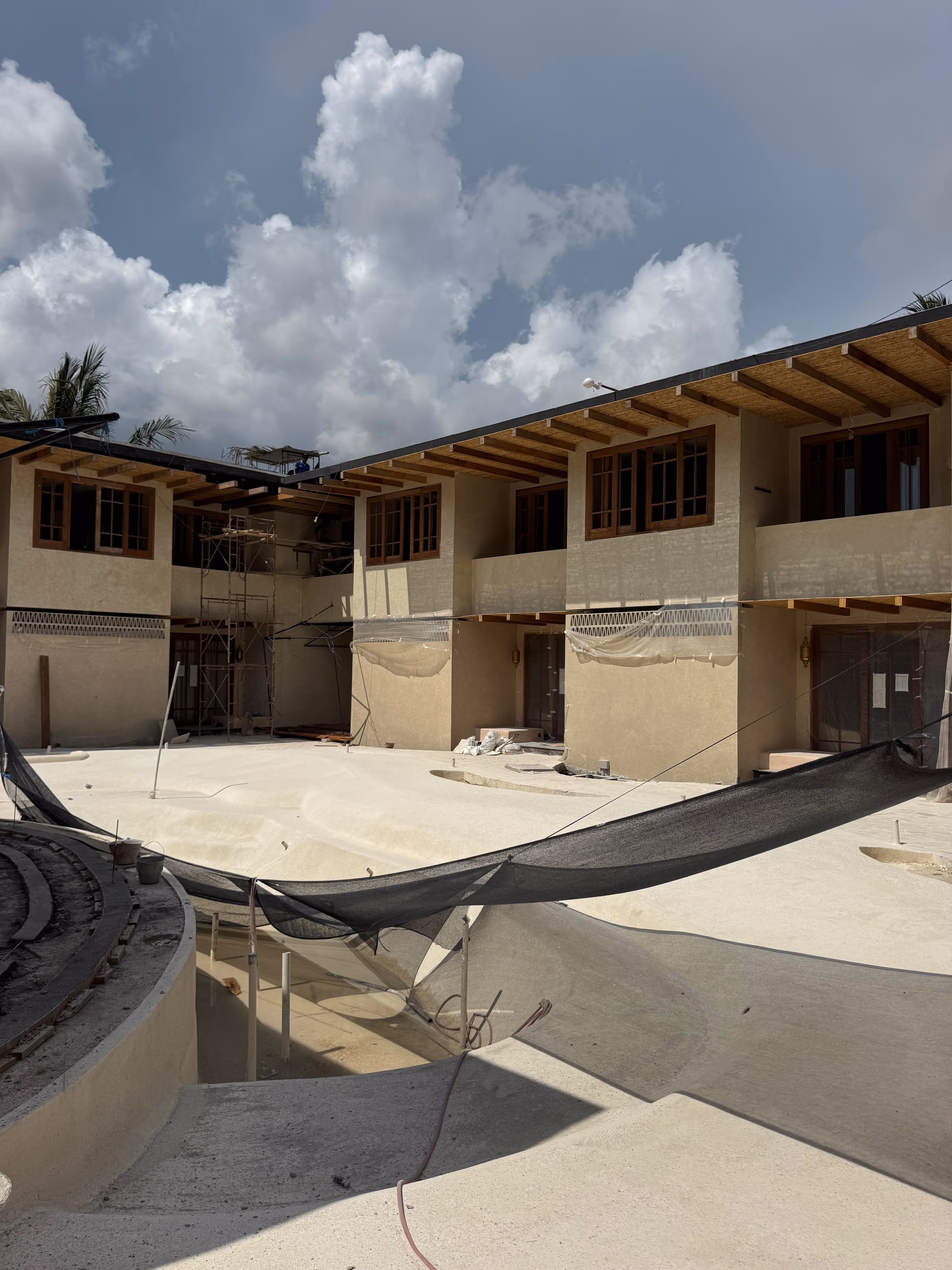 Modern building under construction surrounding an empty pool covered with black protective netting under a cloudy sky.