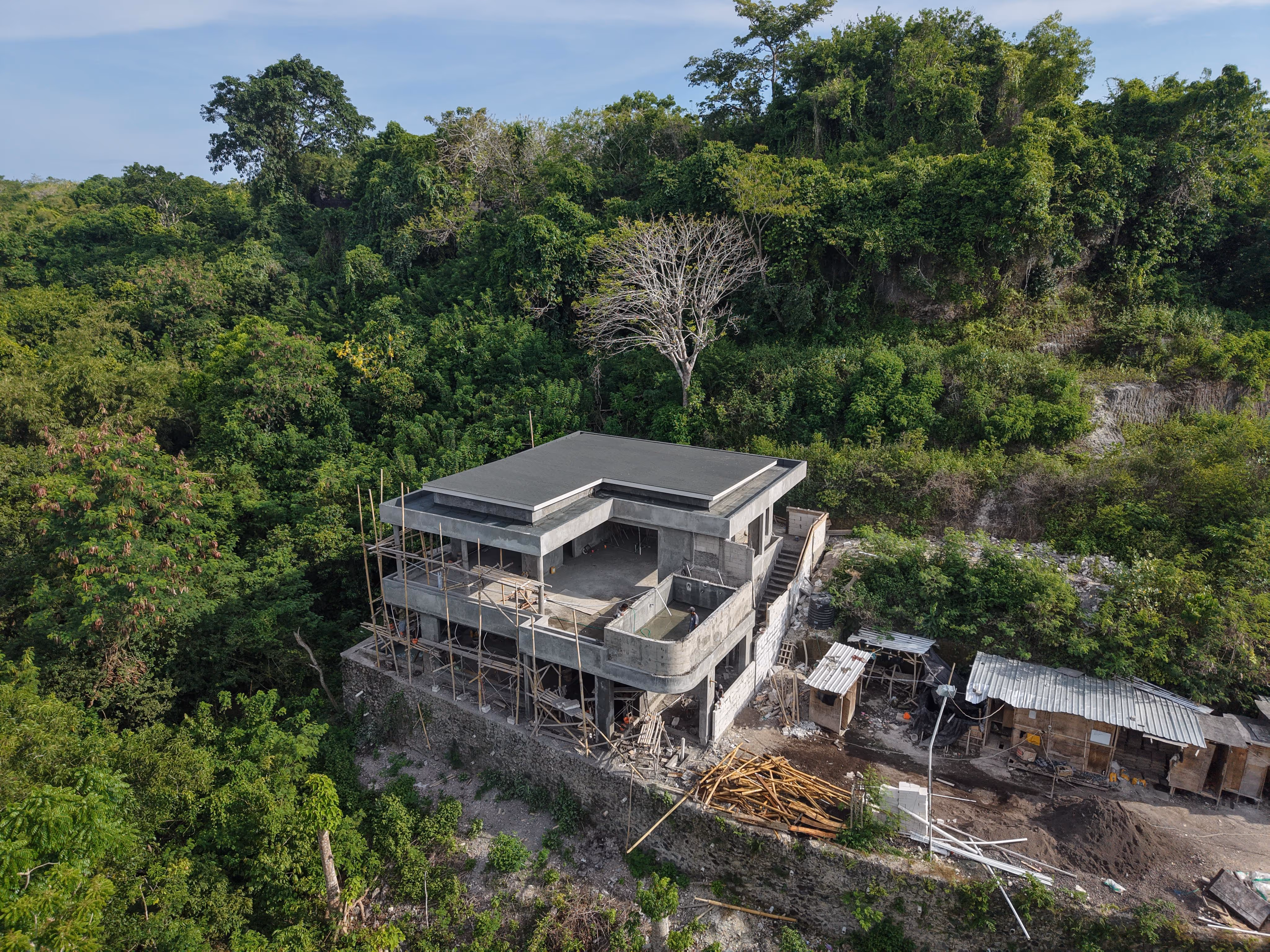 Modern concrete house under construction surrounded by dense green forest and scaffolding on the lower floor.