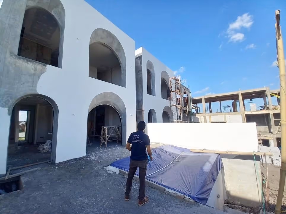 Person standing at a construction site with partially finished white buildings featuring large arched windows and doorways.